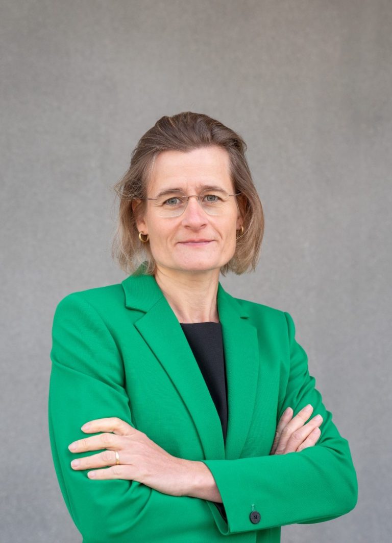 Die Unternehmerin steht mit verschränkten Armen und einem grünen Blazer vor einer grauen Wand.