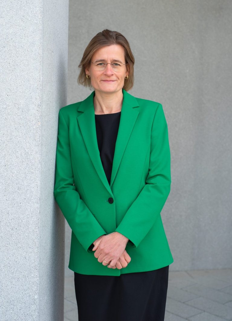 Die Unternehmerin steht mit verschränkten Armen und einem grünen Blazer vor einer grauen Wand.